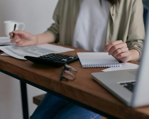 A woman manages finances at home, using a laptop and calculator on a wooden desk.